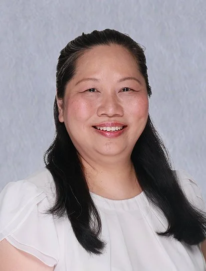 Woman with short dark hair, smiling, wearing a blue blazer over a white shirt, standing against a plain white wall.
