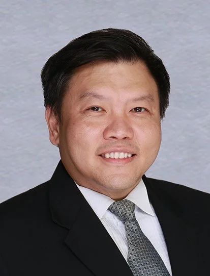 A man in a black suit with a blue shirt and a patterned tie, crossing his arms and wearing a silver wristwatch, posing against a plain white background.