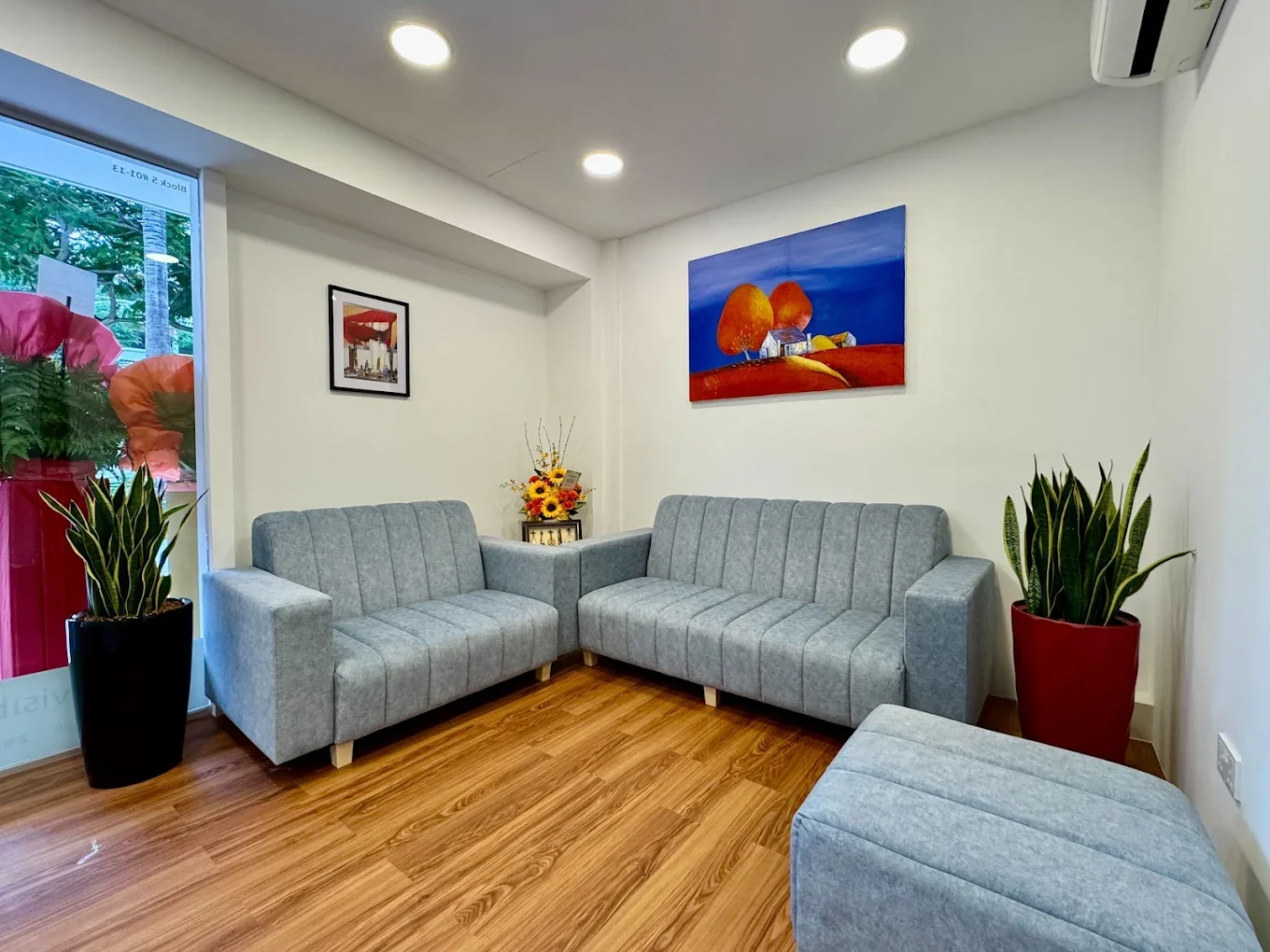 A cozy waiting room with two gray sofas arranged in an L-shape, a small gray ottoman in the foreground, and a vase with yellow flowers on a side table. There's a large window with pink and orange flowers outside, and the room features two framed wall