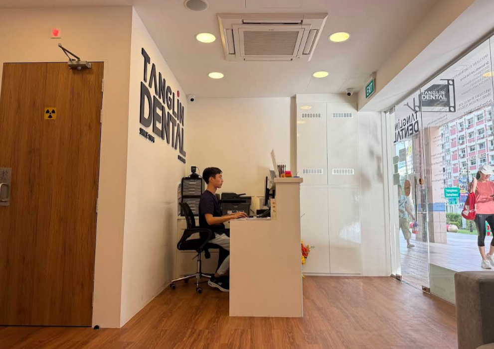 Reception area of a dental clinic with a young man sitting at a desk working on a computer. The sign on the wall reads "Tanglin Dental." The front door is made of glass, showing people walking outside.