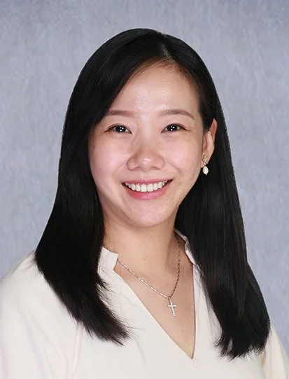 A portrait of a woman with long black hair, smiling, wearing pearl earrings and a dark blue button-up shirt.