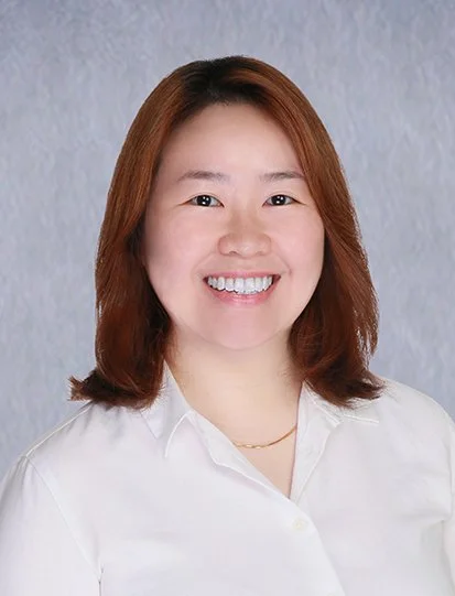 A woman with short dark hair smiling, wearing a navy blue shirt with small white polka dots, standing in front of a white background.