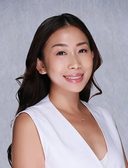 A young woman with long dark hair smiling, wearing a white blazer over a white top, standing against a neutral background.