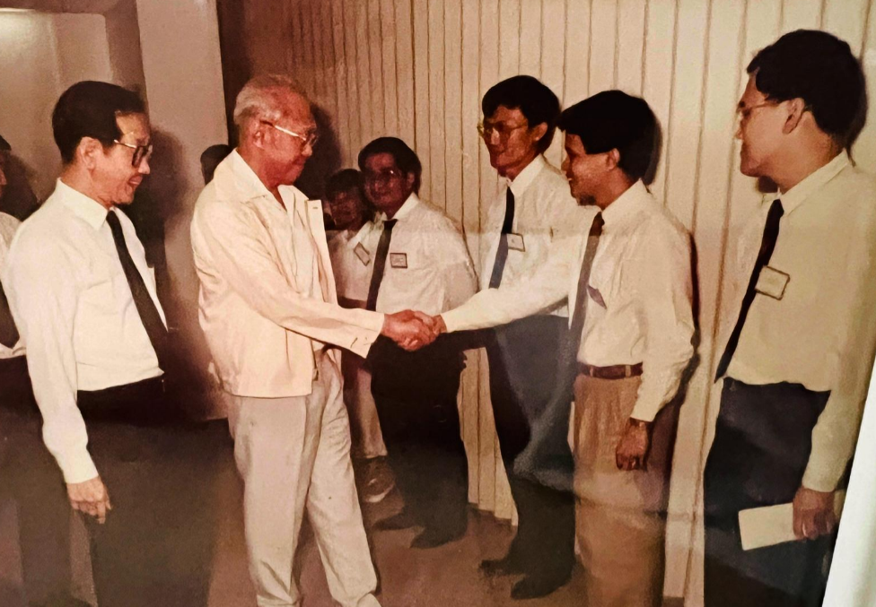 A man with white hair and glasses wearing a cream-colored suit shaking hands with a younger man in a white shirt and black tie, surrounded by other people dressed similarly, in an indoor setting.
