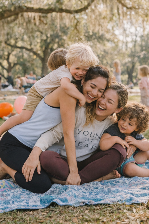 two moms sitting down laughing with their children crawling all over them