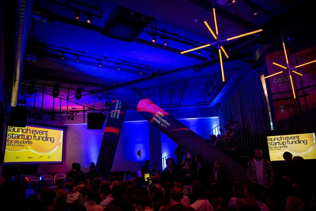 Selskapslokalet Hallen hos Trekanten, dekorert med stemningslys, logo projisert på stolper og to skjermer med navnet på et event.