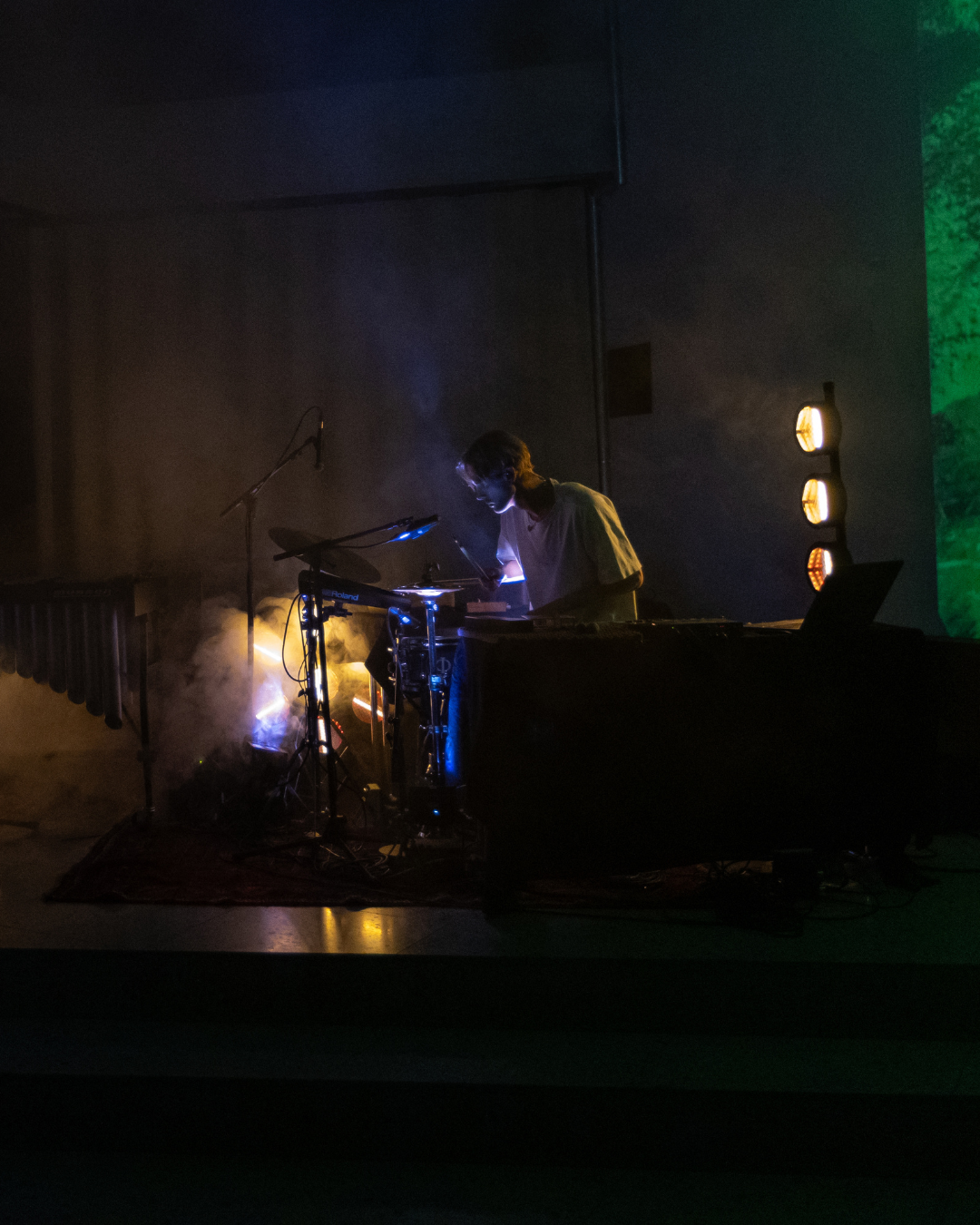 Artist som kombinerer trommer og elektronikk under Ultima x Trekanten, opplyst av blått og gult scenelys i et mørkt konsertrom.