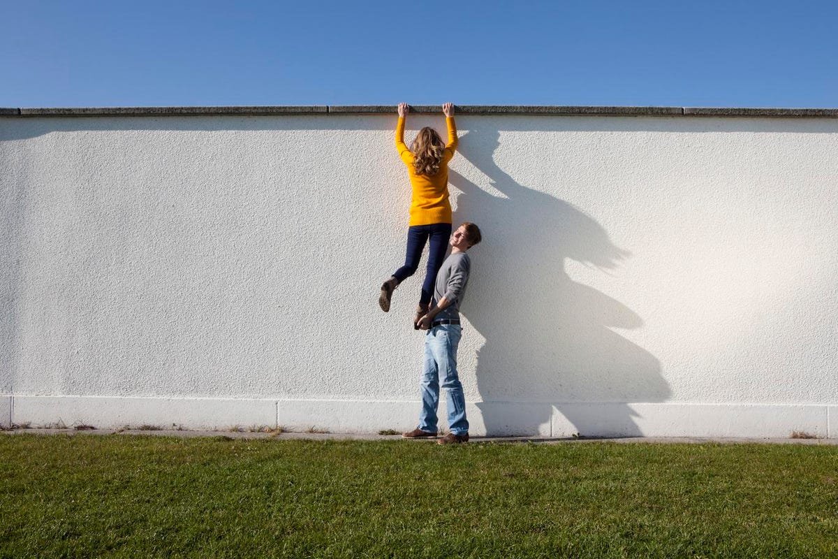 Eine Person, die auf dem Boden neben einer hohen Mauer steht, versucht, einer anderen Person zu helfen, indem sie sie hochschiebt.