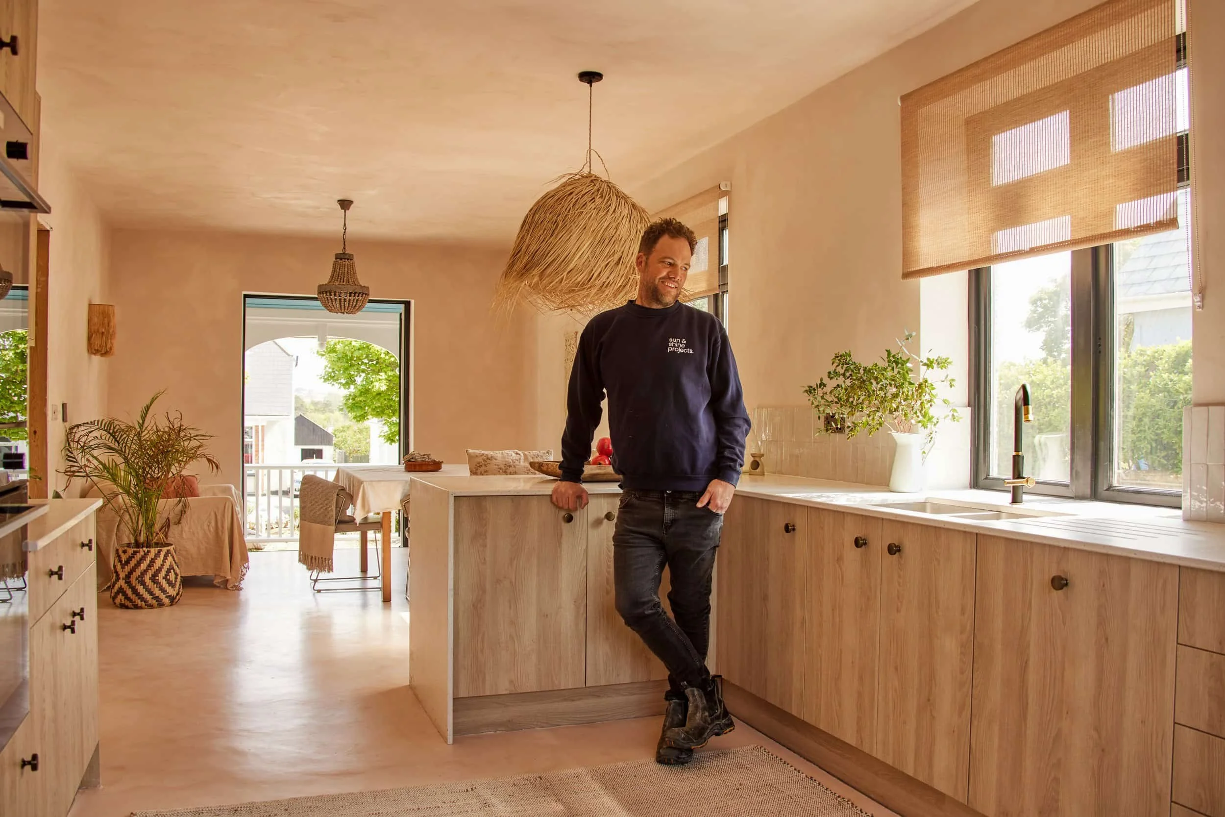 A man standing in a bright, modern kitchen with wooden cabinets, large windows, and a dining area in the background.