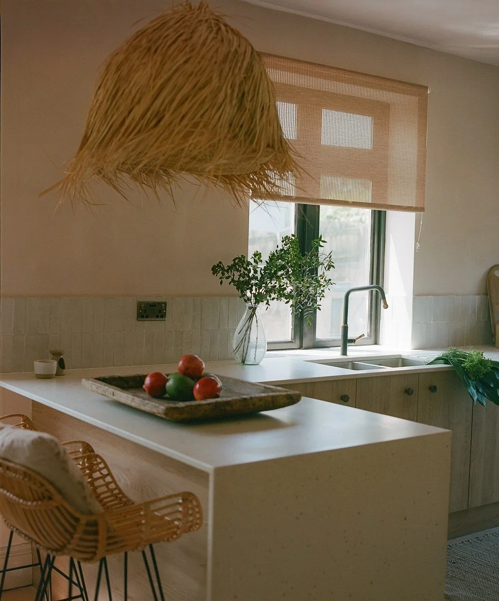 A modern kitchen with a white countertop, a wooden bar stool, a hanging woven lampshade, a vase with green leafy plant, a window with a beige roller shade, a sink with a faucet, and a bunch of leafy greens on the side.