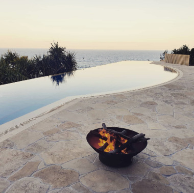 A view of an infinity pool overlooking the ocean at sunset, with a fire pit on a stone patio in the foreground.