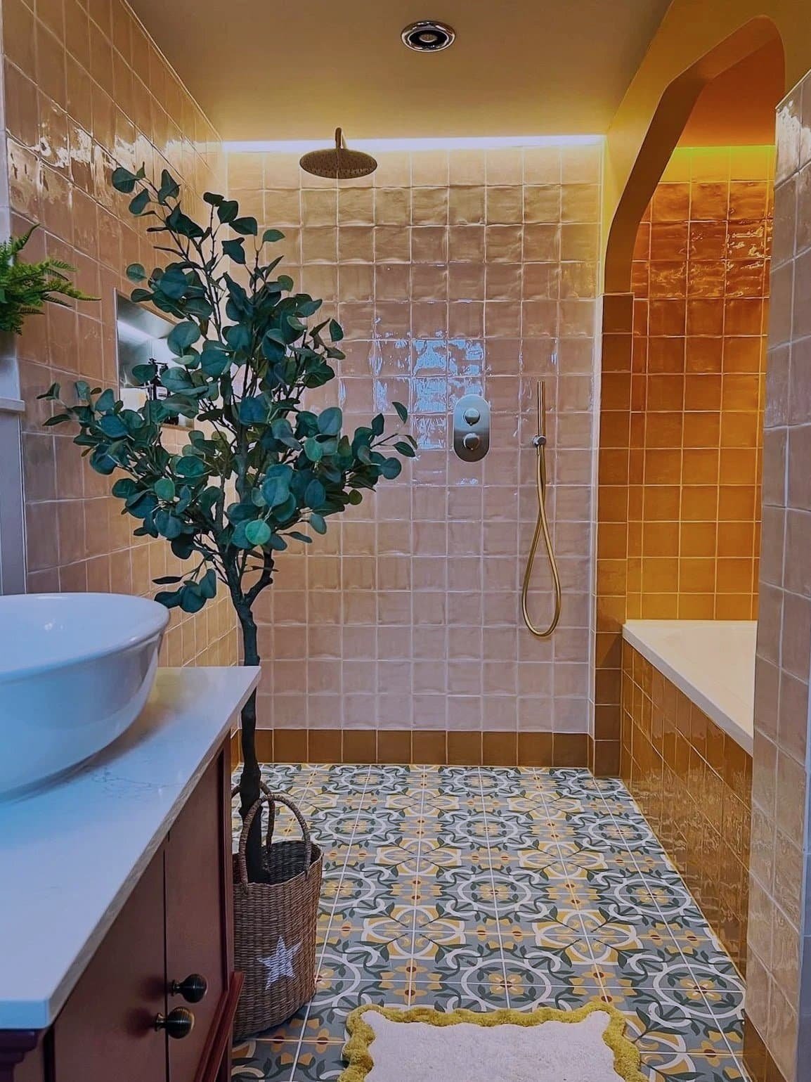 A bathroom with peach-colored tiled walls and a multicolored patterned tile floor. It features a large indoor plant in a basket, a white vessel sink on a wooden cabinet, and a walk-in shower with a rainfall showerhead and a handheld shower, along with a built-in bathtub. There is a yellow and white rug on the floor.
