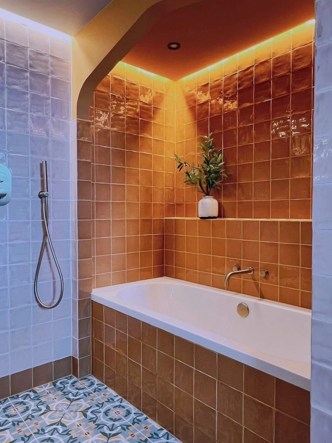 Modern bathroom with a built-in bathtub, a plant in a white pot placed on a tiled shelf above the tub, orange and white tiled walls, decorative patterned floor tiles, and a ceiling with warm lighting.