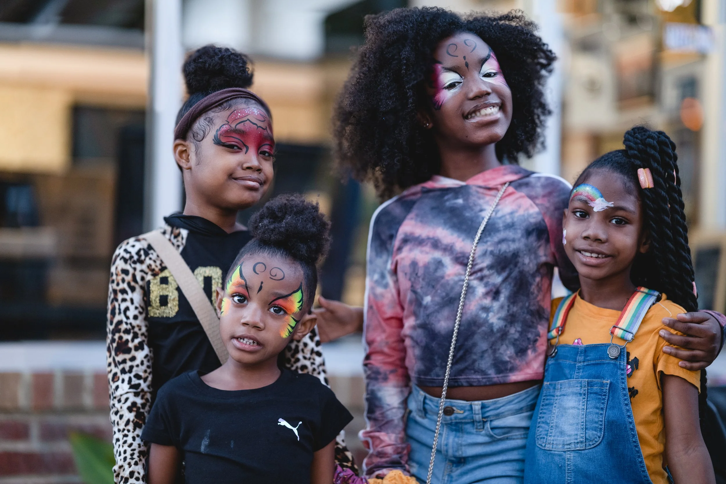 Group of children and young woman with face paint, smiling outdoors in front of a building.