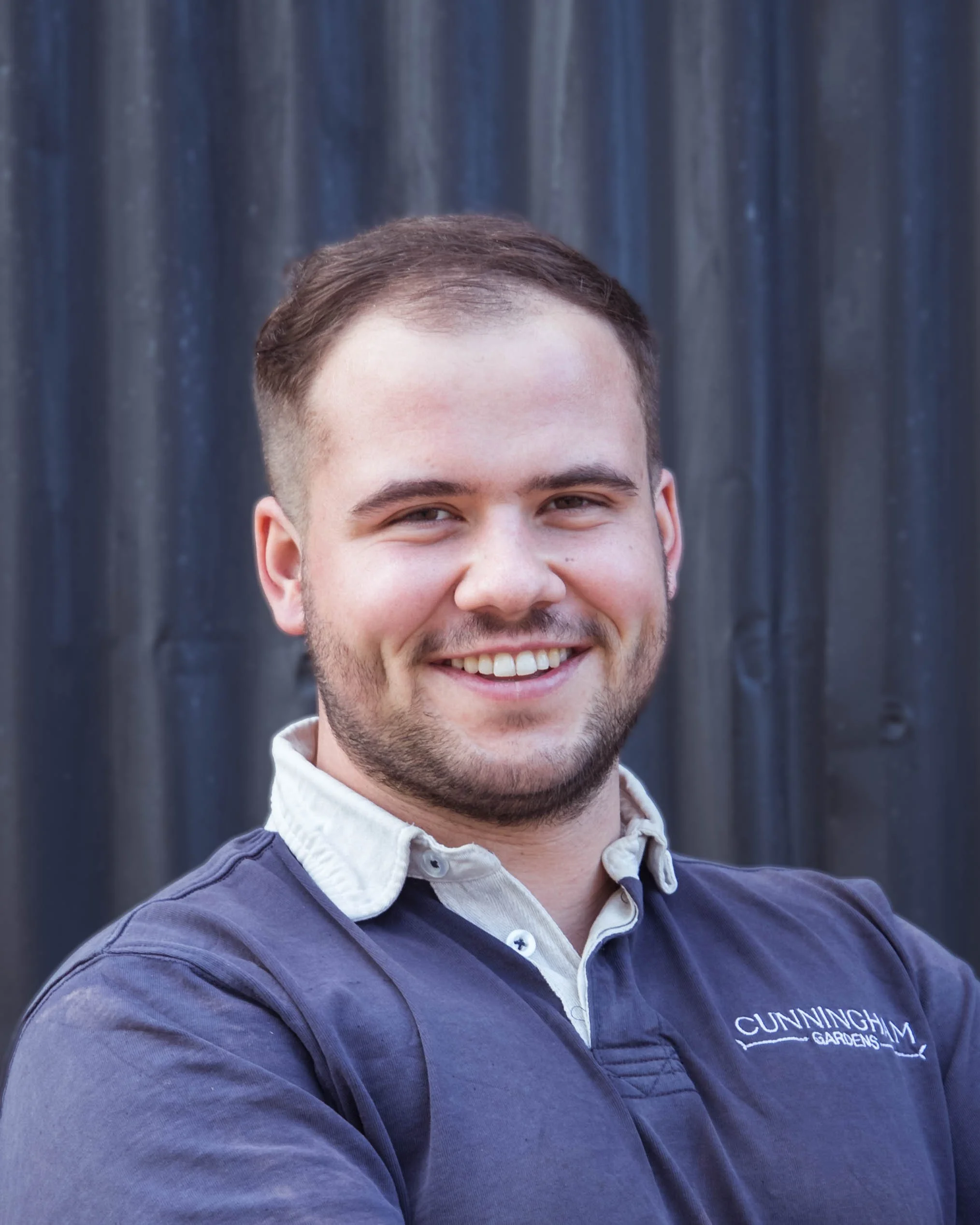 A smiling young man with short brown hair and a beard, wearing a dark blue polo shirt with a white collar, standing outdoors in front of a dark, corrugated metal wall.