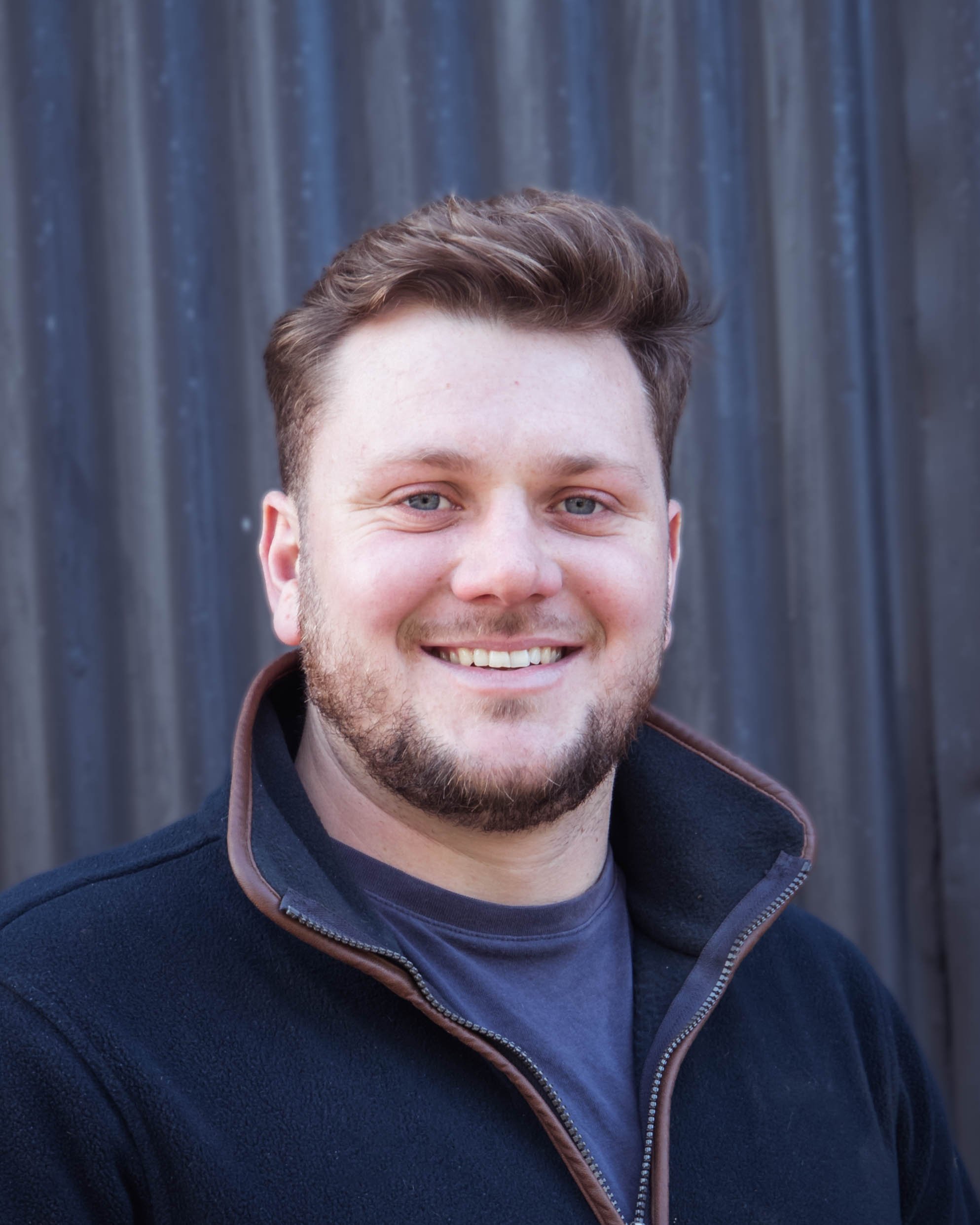 A smiling young man with short brown hair, a beard, and blue eyes, wearing a dark jacket with a zipper, standing in front of a gray, textured background.