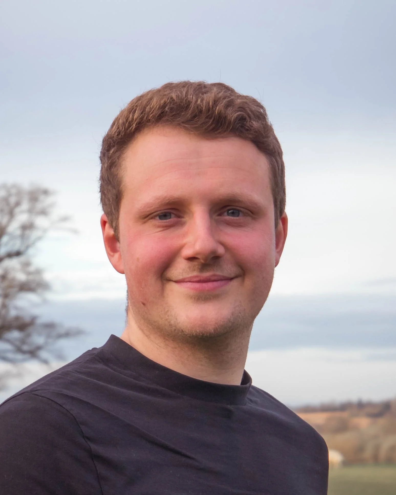 A young man with short brown hair and blue eyes is smiling outdoors, wearing a black shirt. The background features a cloudy sky, trees, and an open landscape.