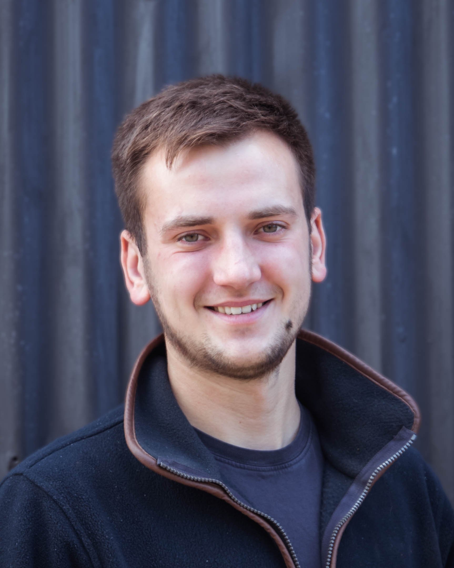 A young man with short brown hair and a light beard smiling, wearing a black jacket with a brown collar, standing in front of a dark, textured background.
