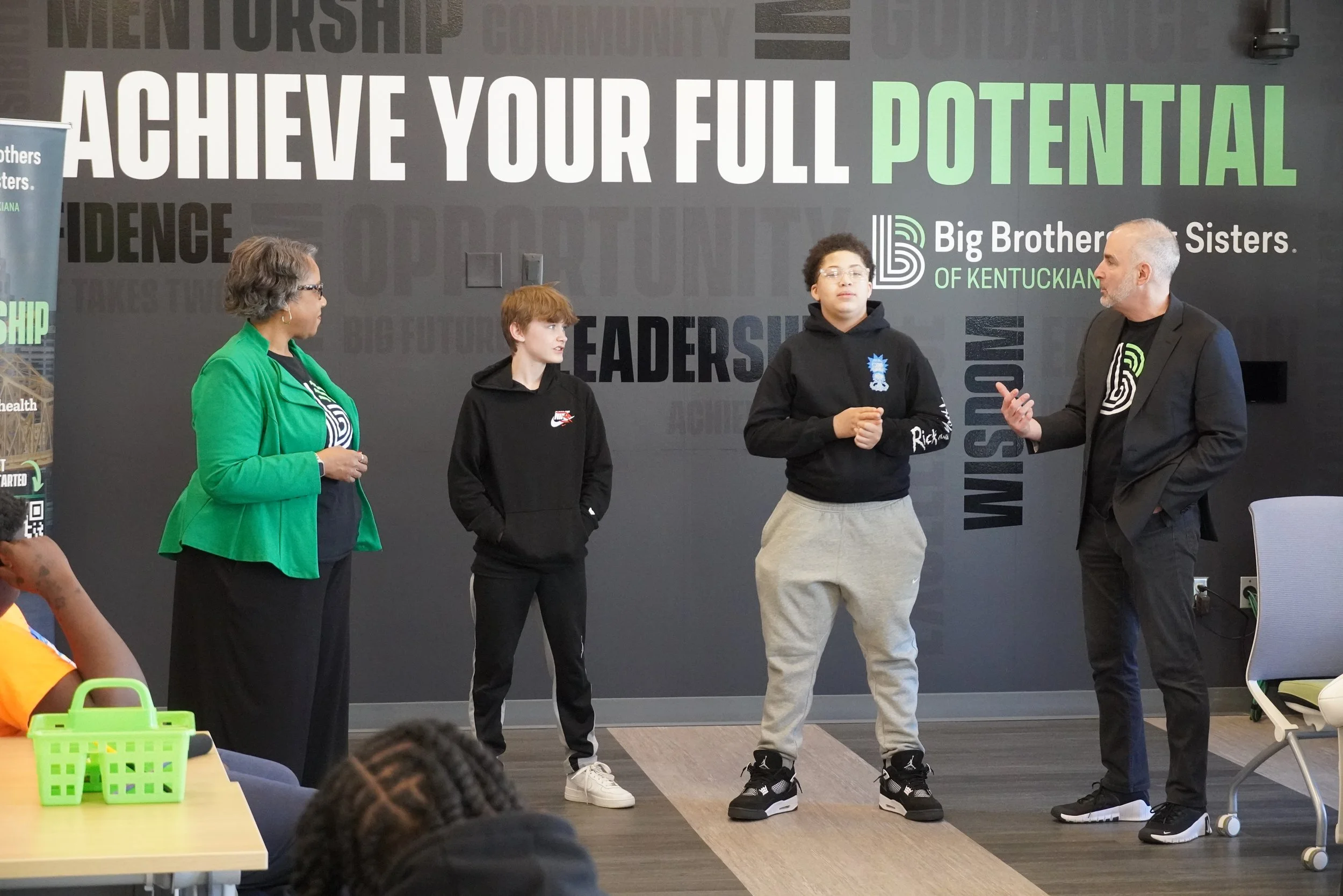 Two young boys, one caucasian and one african american, speak in front of a group with a while male CEO asking them questions in front of a black wall with striking text reading "ACHIEVE YOUR FULL POTENTIAL" in bold text