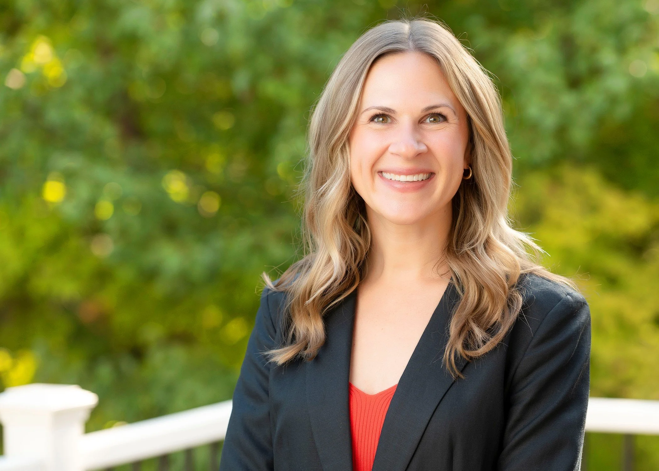 blonde-haired woman smiles while wearing green earrings and a professional blazer outside on a sunny day