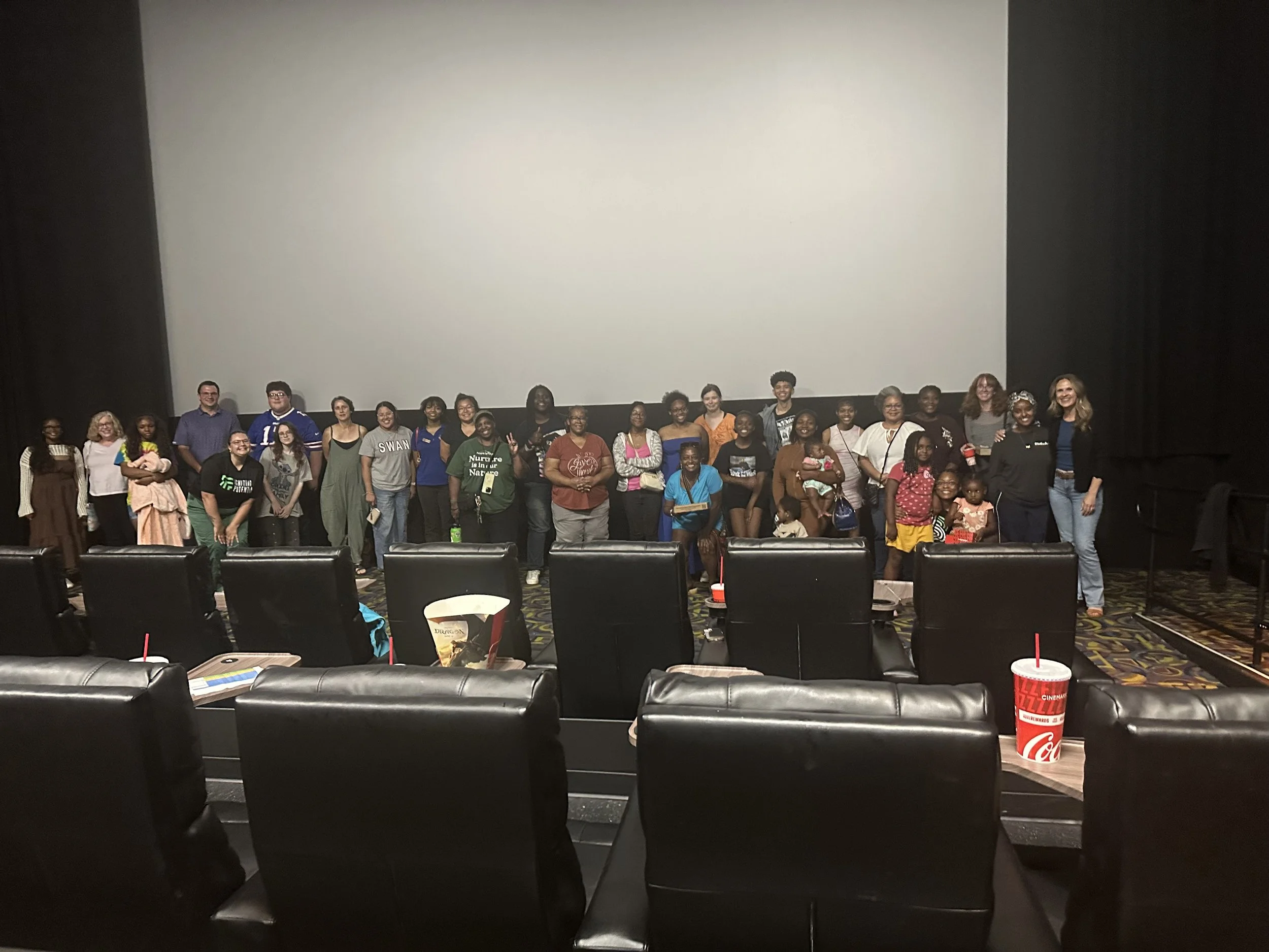 Group of people standing together in a movie theater, facing the camera with chairs and drinks in front.