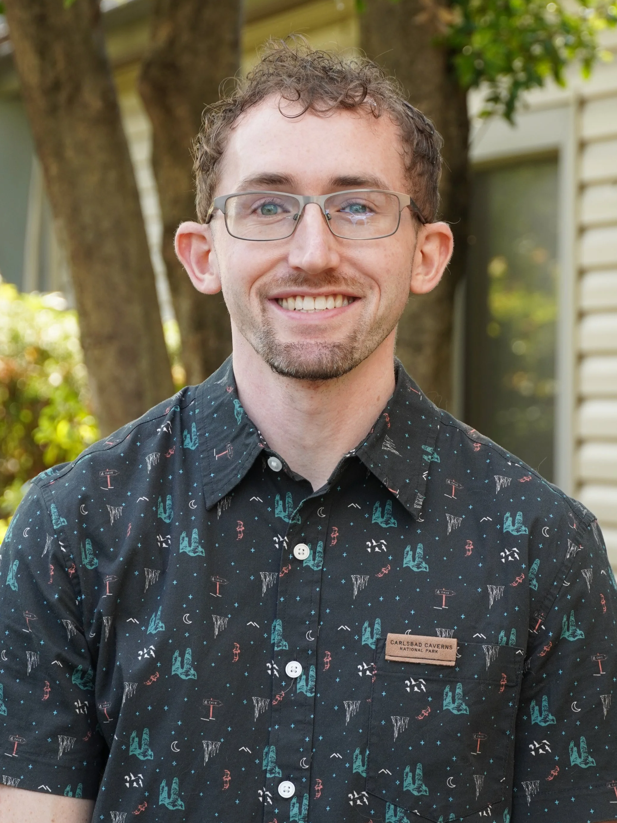 A man with curly hair and glasses stands smiling outside on a sunny day