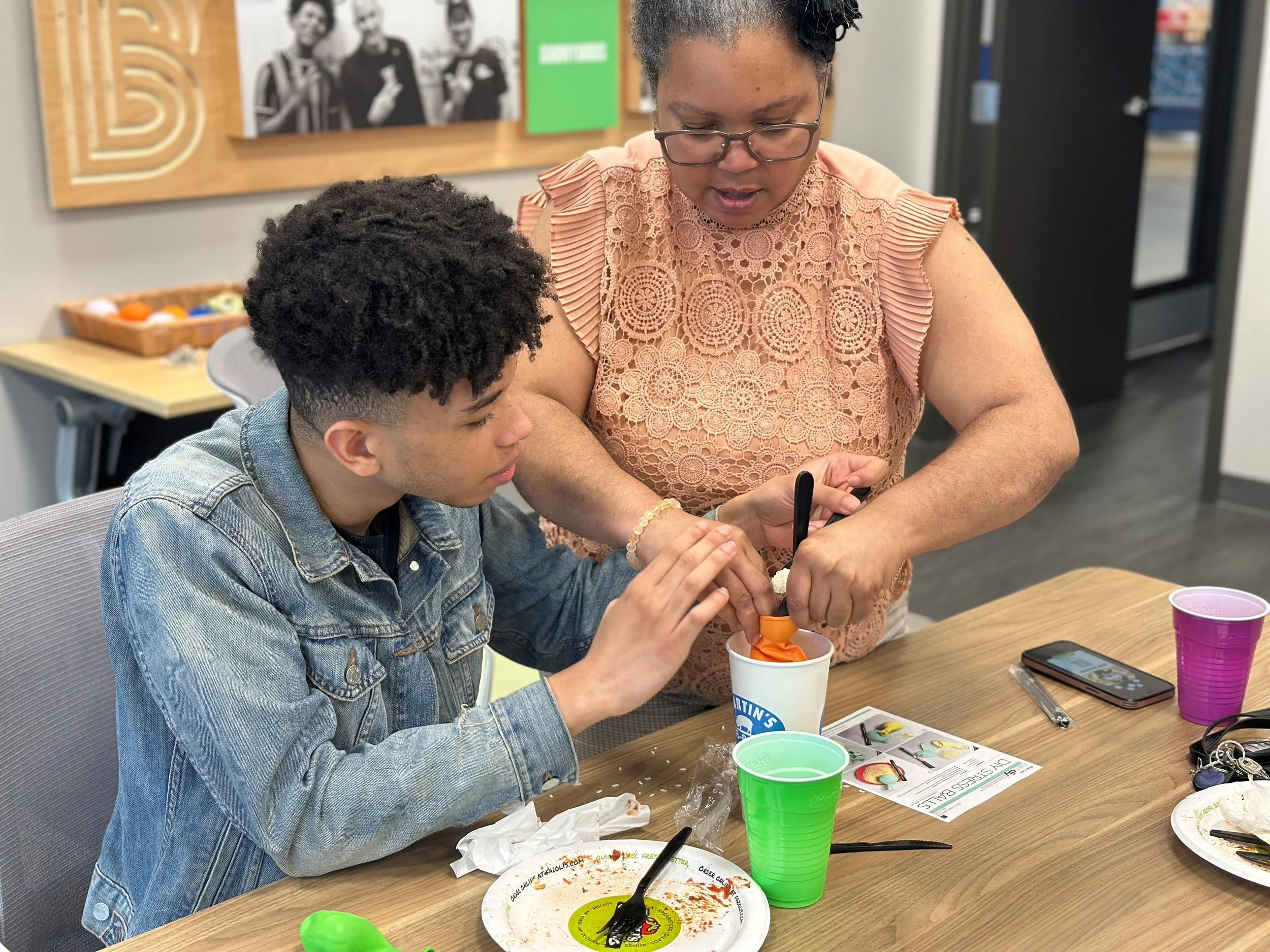 A young man and an older woman are sitting at a table engaged in planting a small plant into a cup together. The table has plates, cups, and utensils on it, and the background shows a bulletin board with photos and decorations.