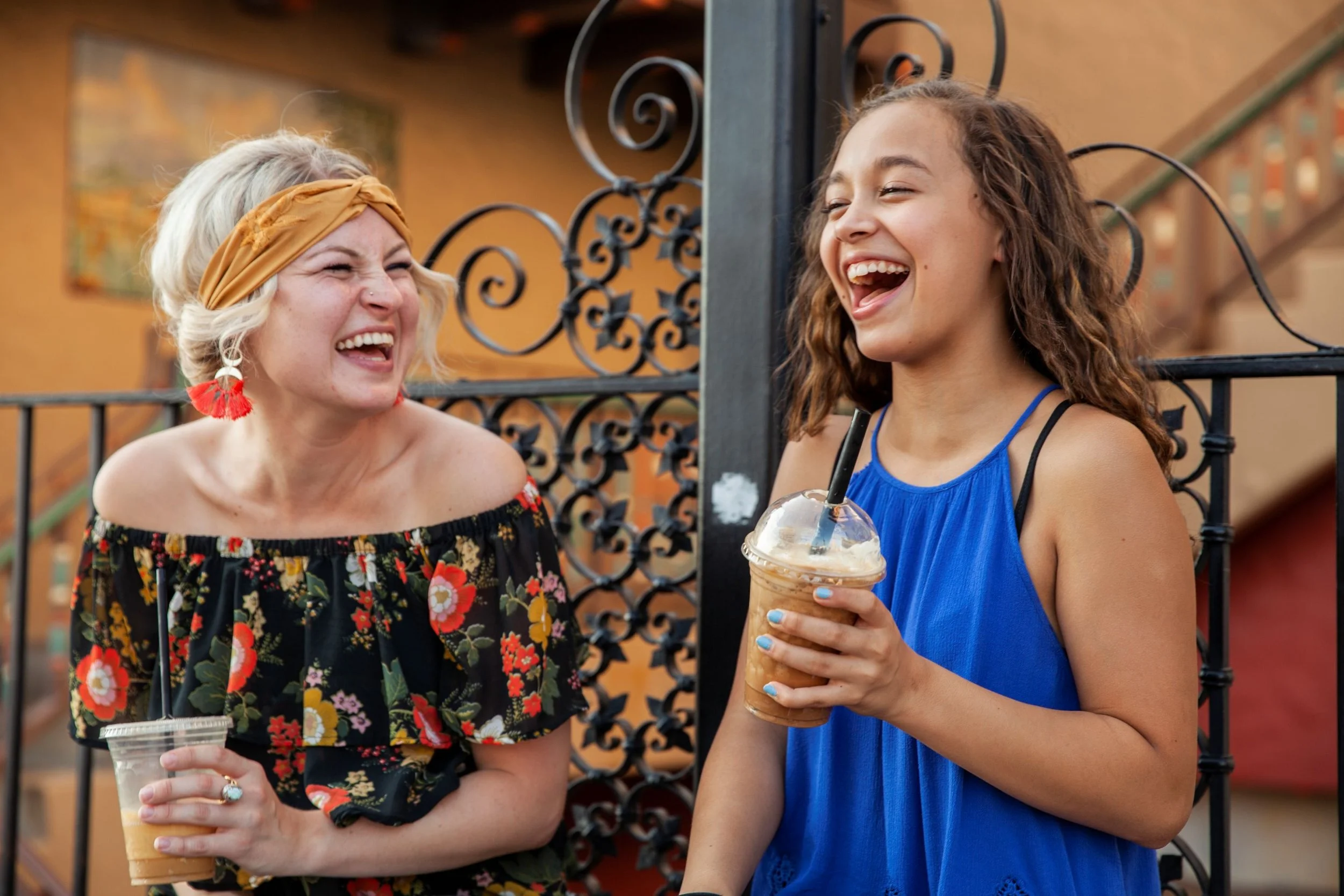 A caucasian blond woman smiles while holding an iced coffee and laughing while sitting next to her mentee, a young mixed race girl wearing a blue top and holding an iced coffee