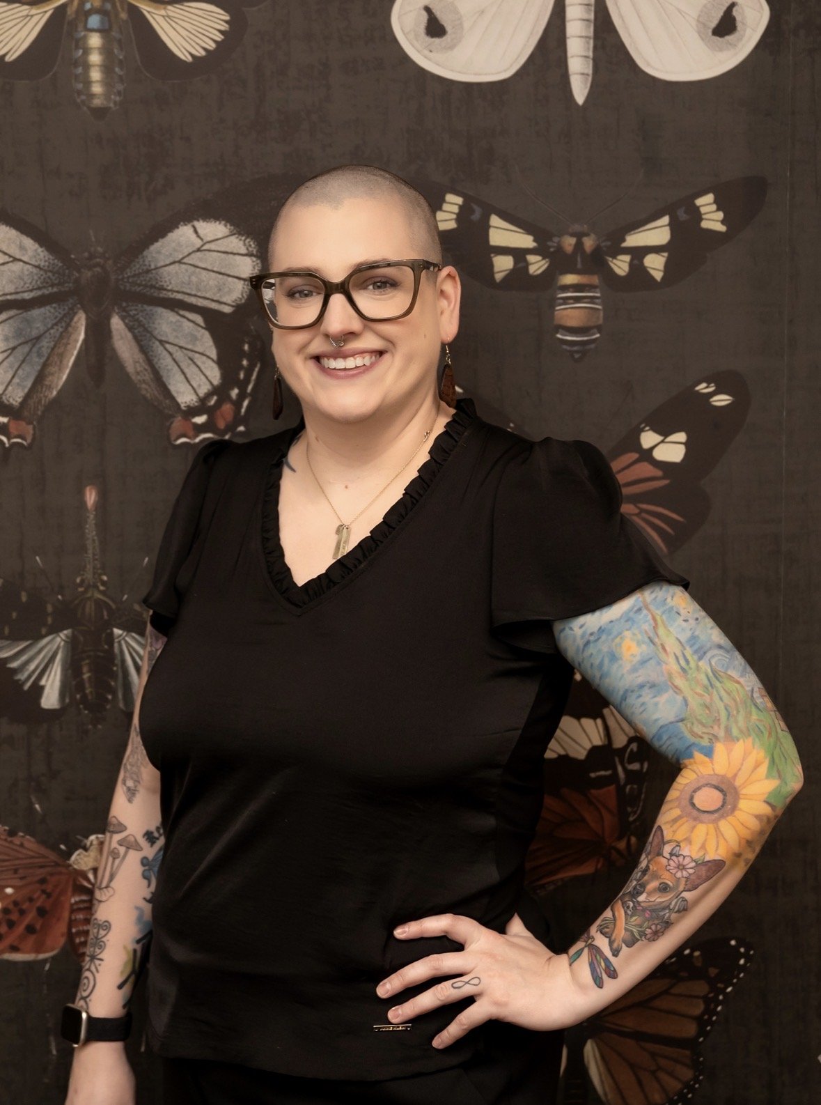 A smiling woman with glasses and a septum piercing, wearing a black top and displaying colorful tattoos on her arms, standing in front of a butterfly-themed wall art.