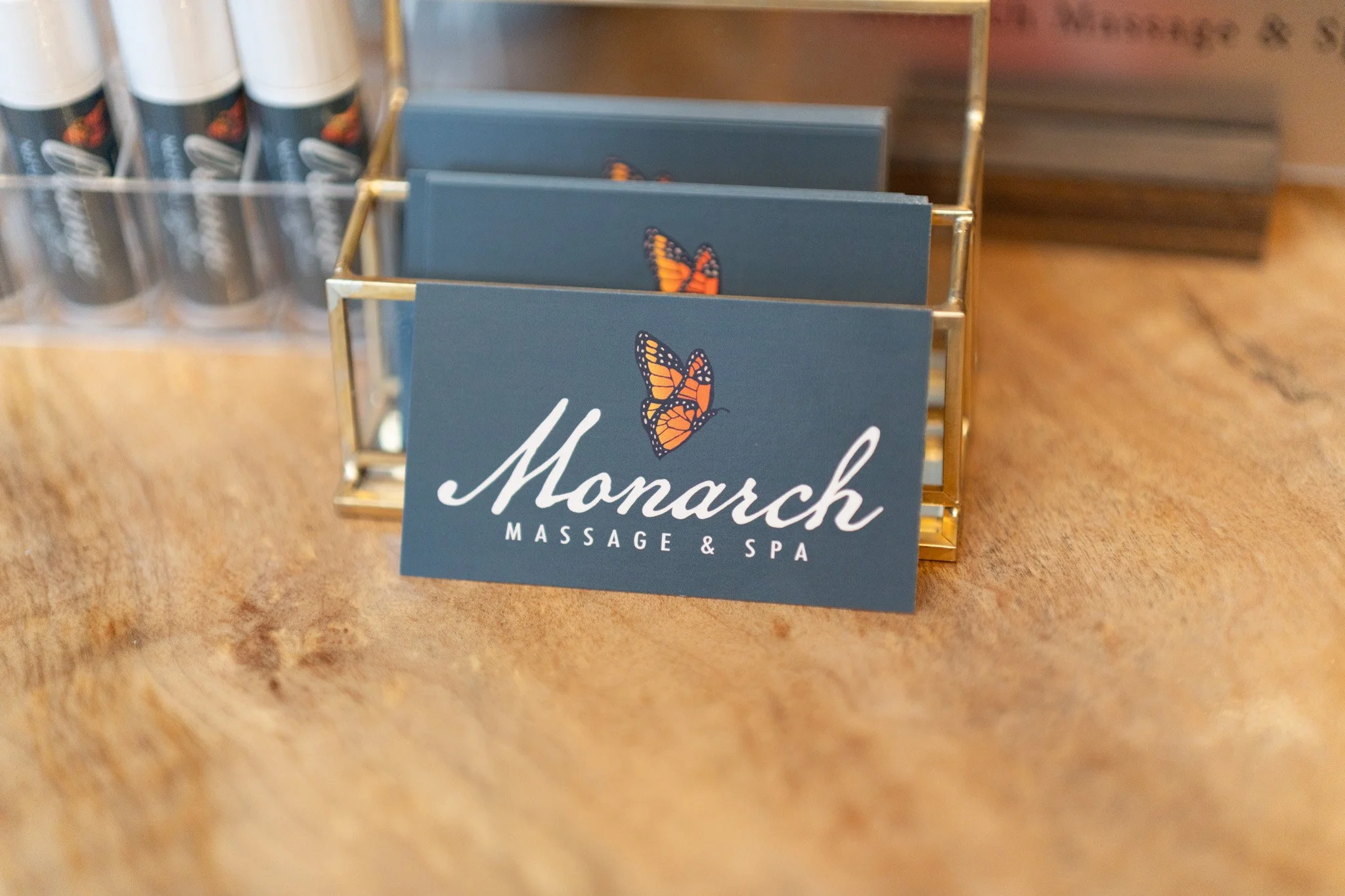 A small sign that reads "Monarch Massage & Spa" with a monarch butterfly illustration, placed on a wooden surface. In the background, there is a display of pamphlets or brochures.