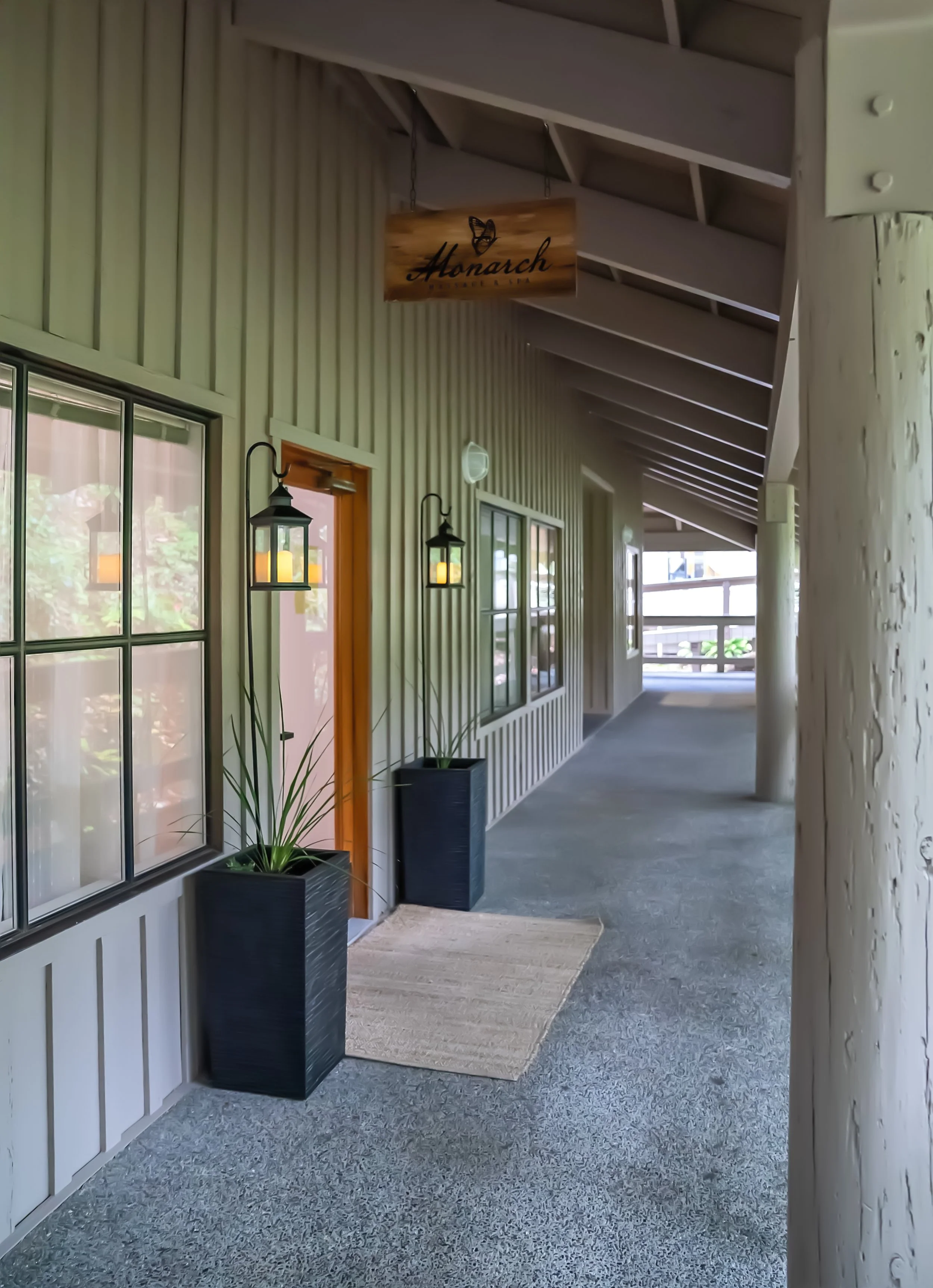 Covered outdoor porch area with vertical beige siding, windows, a door with a wooden frame, potted plants, ceiling lights, and a hanging sign that says 'Monarch'.