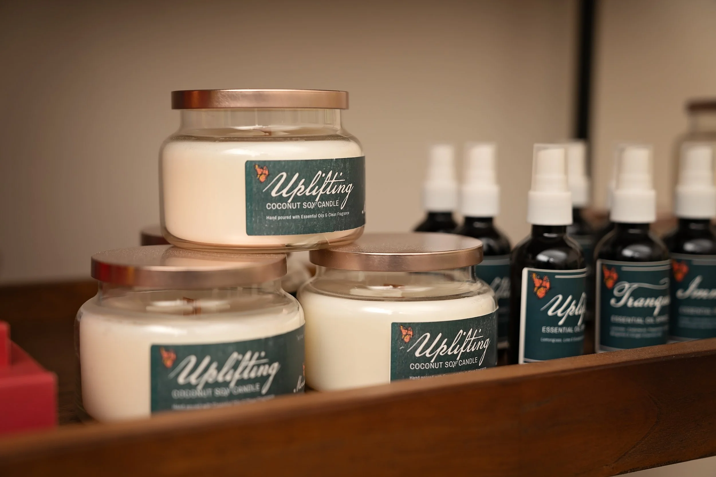 Several candles labeled 'Uplifting Coconut Soy Candle' placed on a wooden shelf, with some dark-colored bottles in the background.