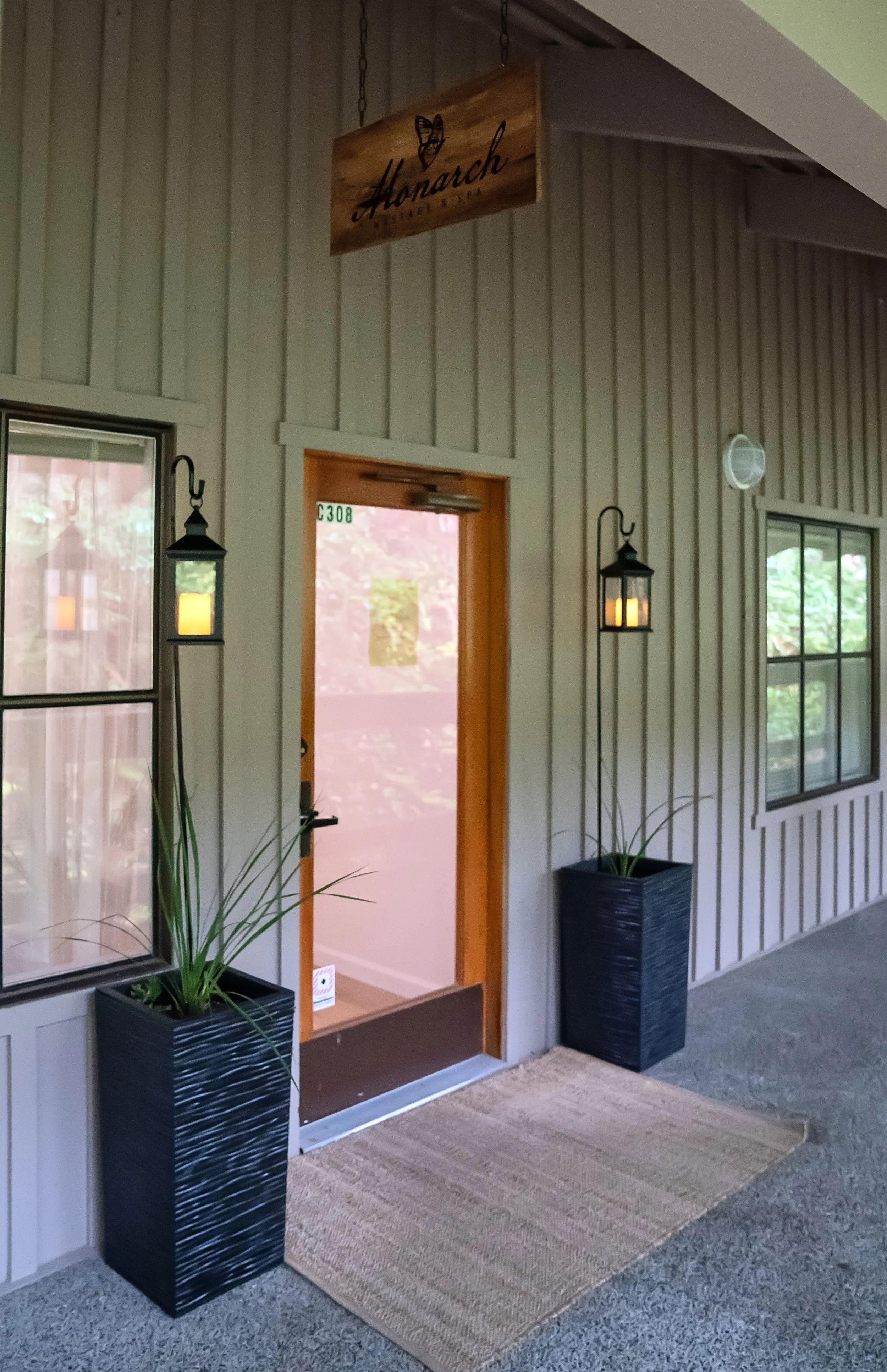 Entryway of a building with wooden sign reading 'The Monarch Ticket & Spa' hanging above the doorway. Two black planters with green plants flank the door, which is partially open. Black lantern-style lights are mounted on the beige vertical wooden paneling. A rug is placed in front of the door on a gray carpeted floor, with windows on each side of the entrance.