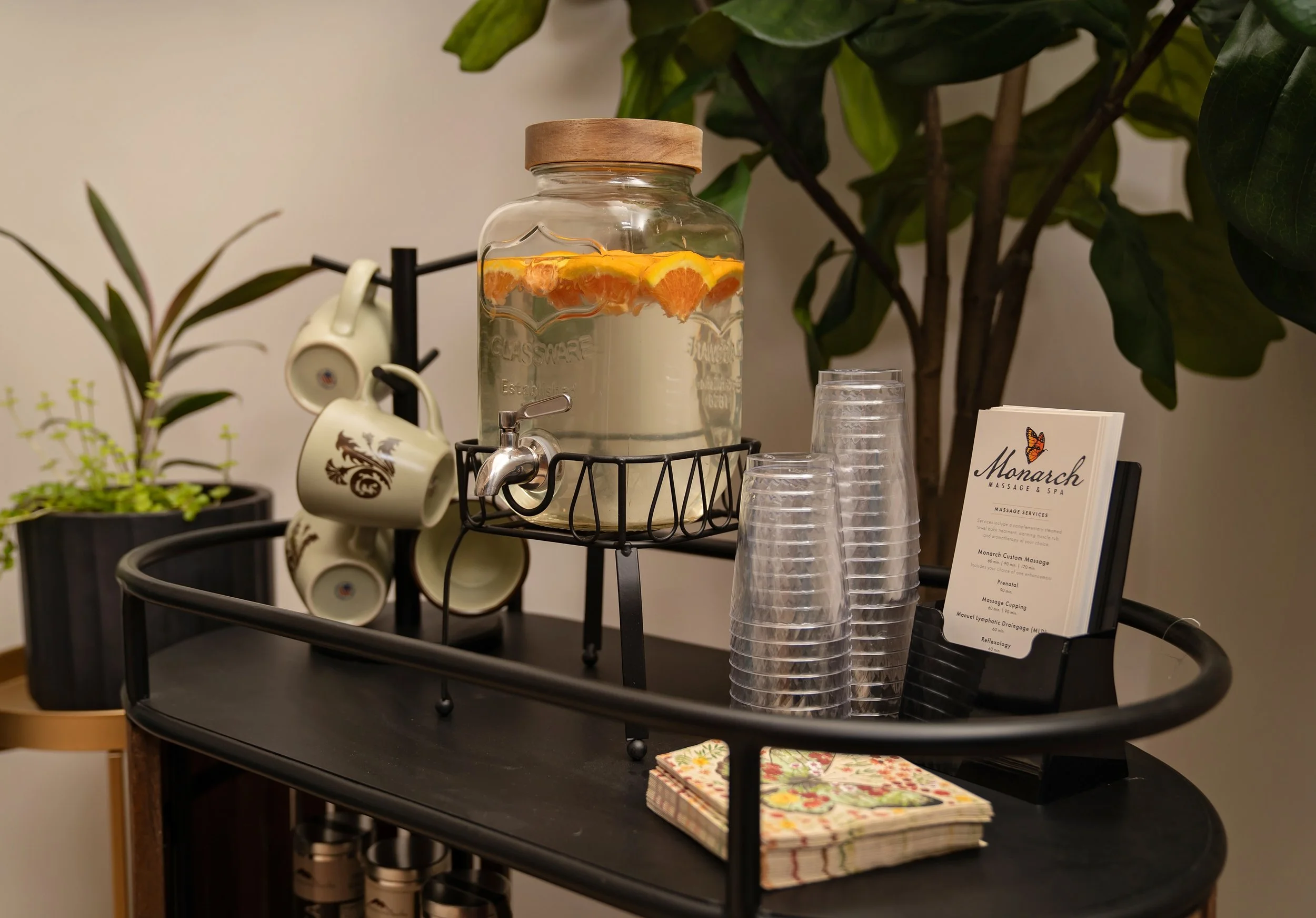 A black three-tiered cart holding a glass jar of sliced oranges in water, clear plastic cups, a small plant, and a printed menu from Monarch Massage & Spa.