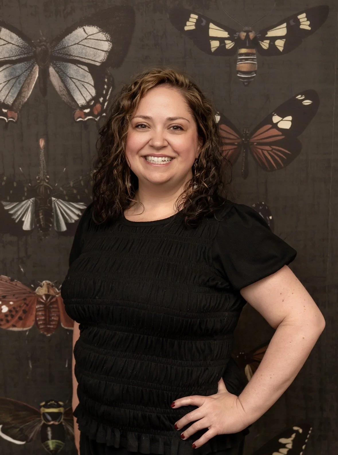 A woman with curly brown hair smiling, wearing a black top, standing in front of a dark background with large butterfly illustrations.