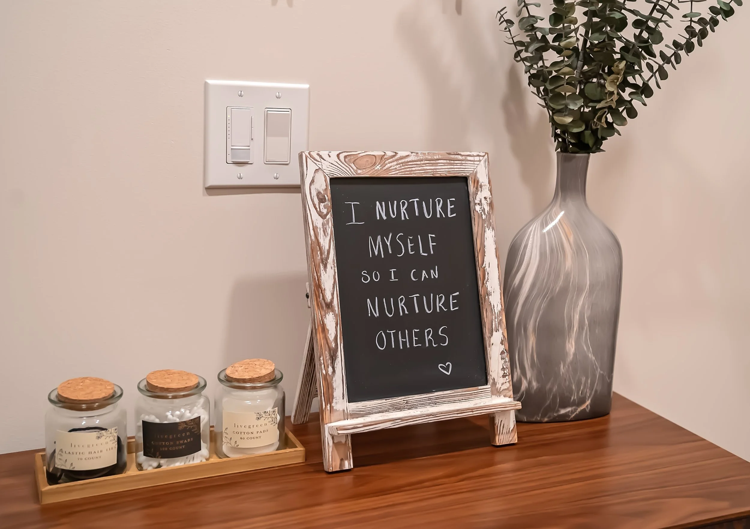 A decorative tabletop setting with three small jars of candles or skincare products, a framed chalkboard sign with a quote about nurturing oneself and others, and a tall vase with green leafy branches.