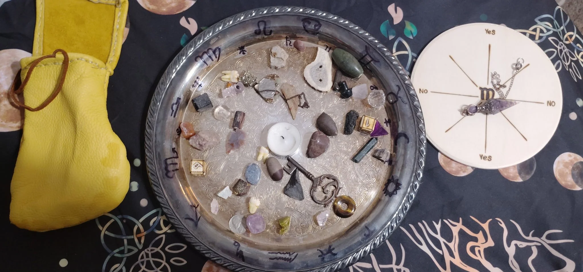 A tarot or spiritual divination setup featuring a silver tray with various gemstones, crystals, and small objects, a white candle, and a round wooden chart with zodiac signs and the word 'Yes' and 'No' for yes/no answers, all on a dark cloth with celestial and abstract patterns, alongside a yellow leather pouch.