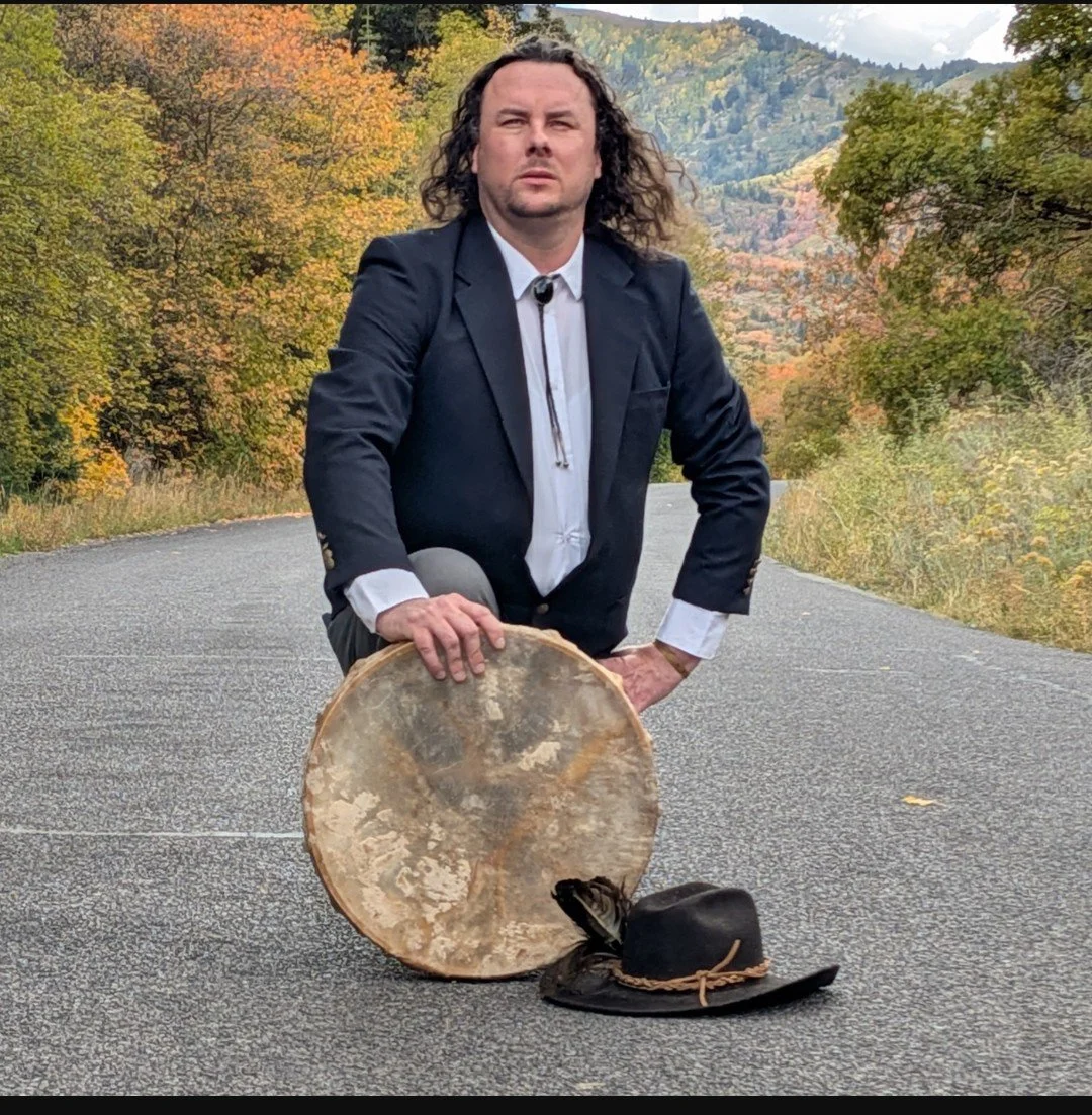 A man in a suit crouching on a deserted road in a mountainous area with autumn-colored trees in the background, resting his hand on a large tree trunk and wearing a cowboy hat placed on the ground nearby.