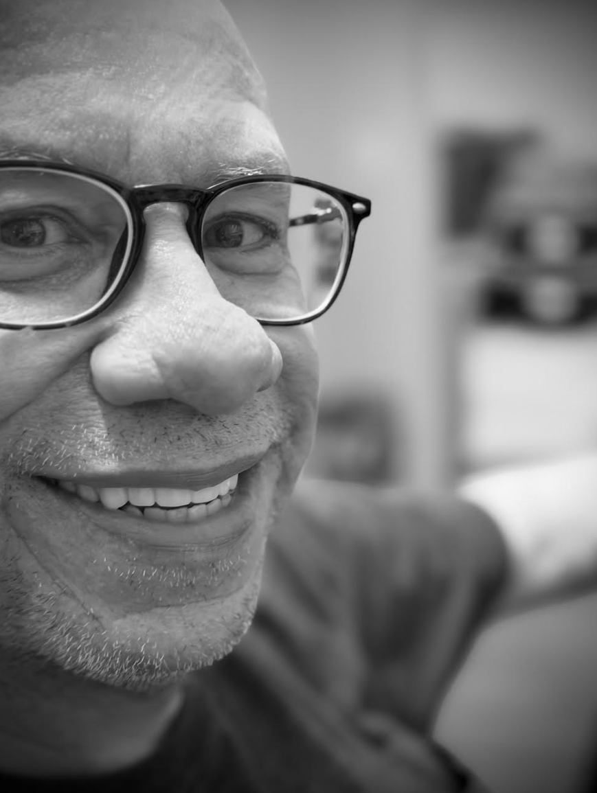 Close-up black and white portrait of smiling man wearing glasses with short facial hair, indoors.