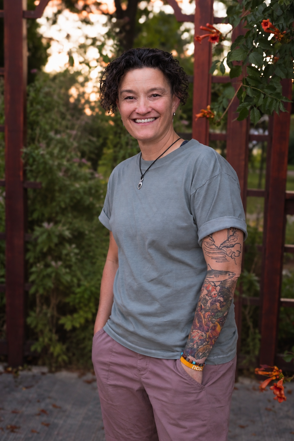 Smiling woman with short curly hair and colorful arm tattoos standing in a flower garden.