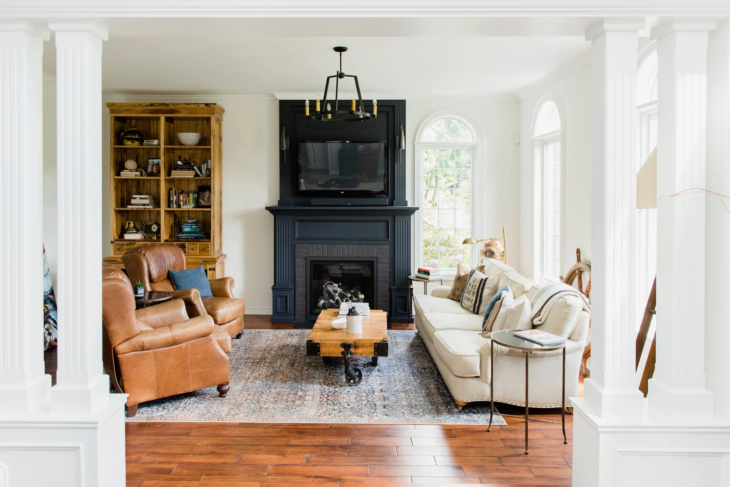 Living room with white walls, hardwood floors, a black fireplace with a mounted TV, beige sofa, leather armchair, wooden bookshelf, and large windows letting in natural light.