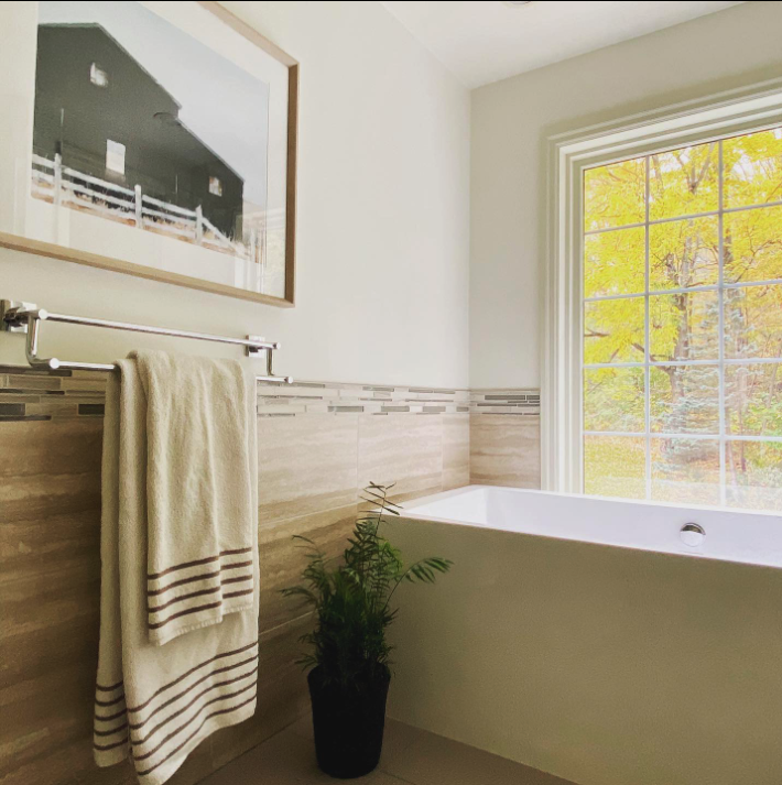 A bathroom with a large window showing trees outside. There is a white bathtub by the window, a towel rack with a beige towel, a framed picture on the wall, and a potted green plant on the floor.