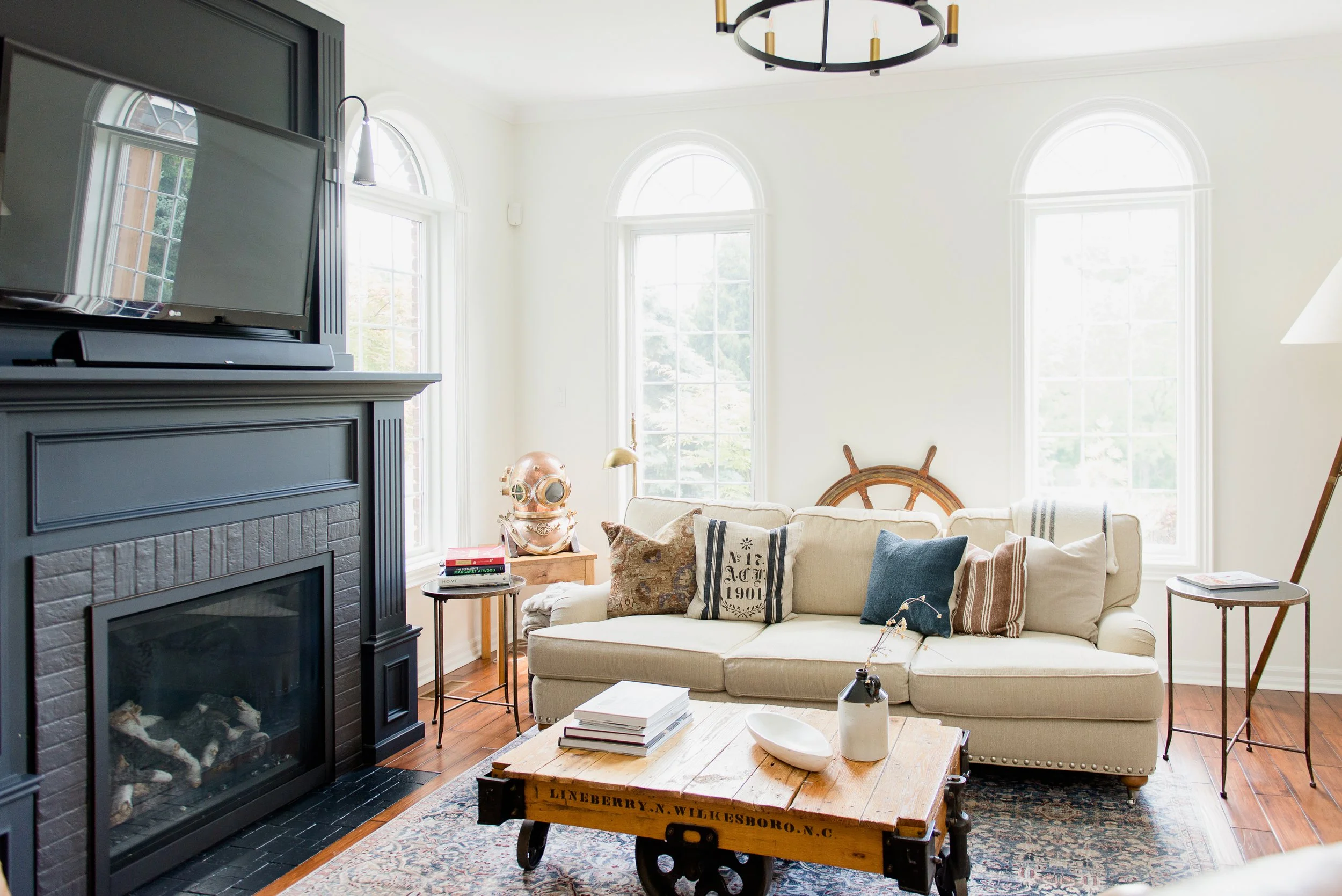 Living room with cream sofa, throw pillows, three large arched windows, wooden coffee table, black fireplace with TV, side tables, decorative items, and hardwood flooring with a patterned rug.