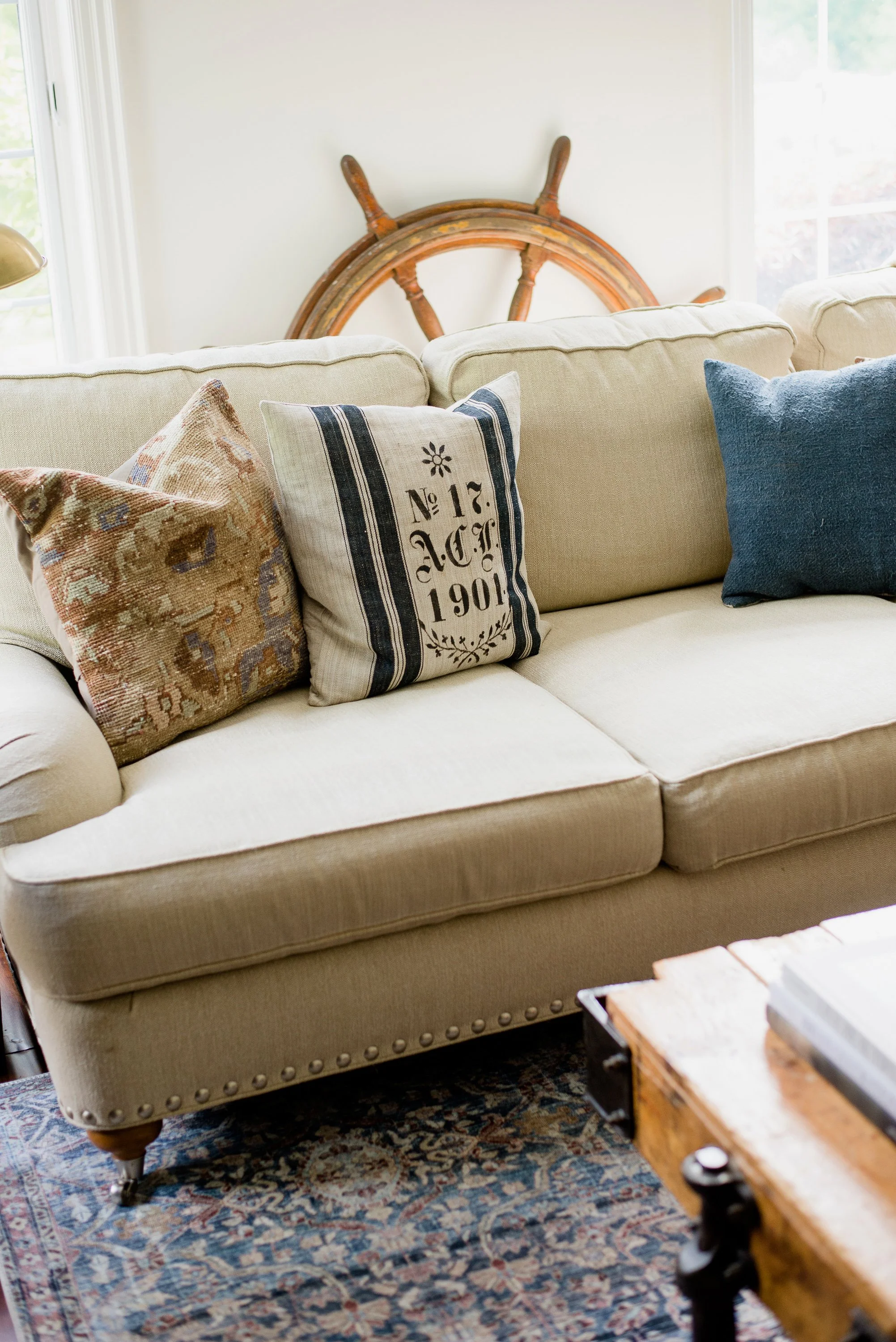 Living room with beige upholstered sofa holding three throw pillows; an old wooden ship's wheel is visible behind the sofa, near a window.