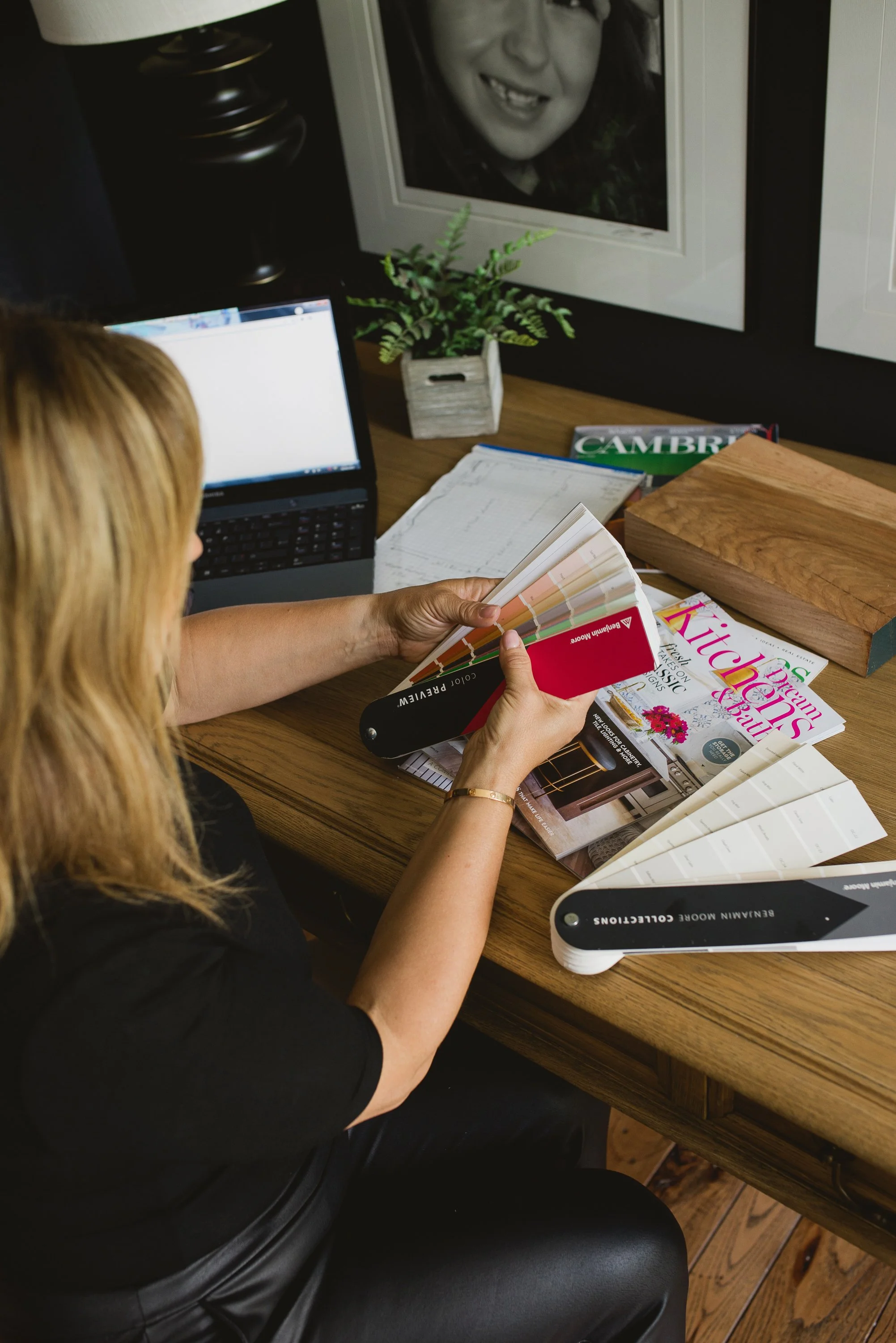 A woman sitting at a wooden desk, looking through paint color samples while surrounded by magazines, a laptop, and a color fan deck.