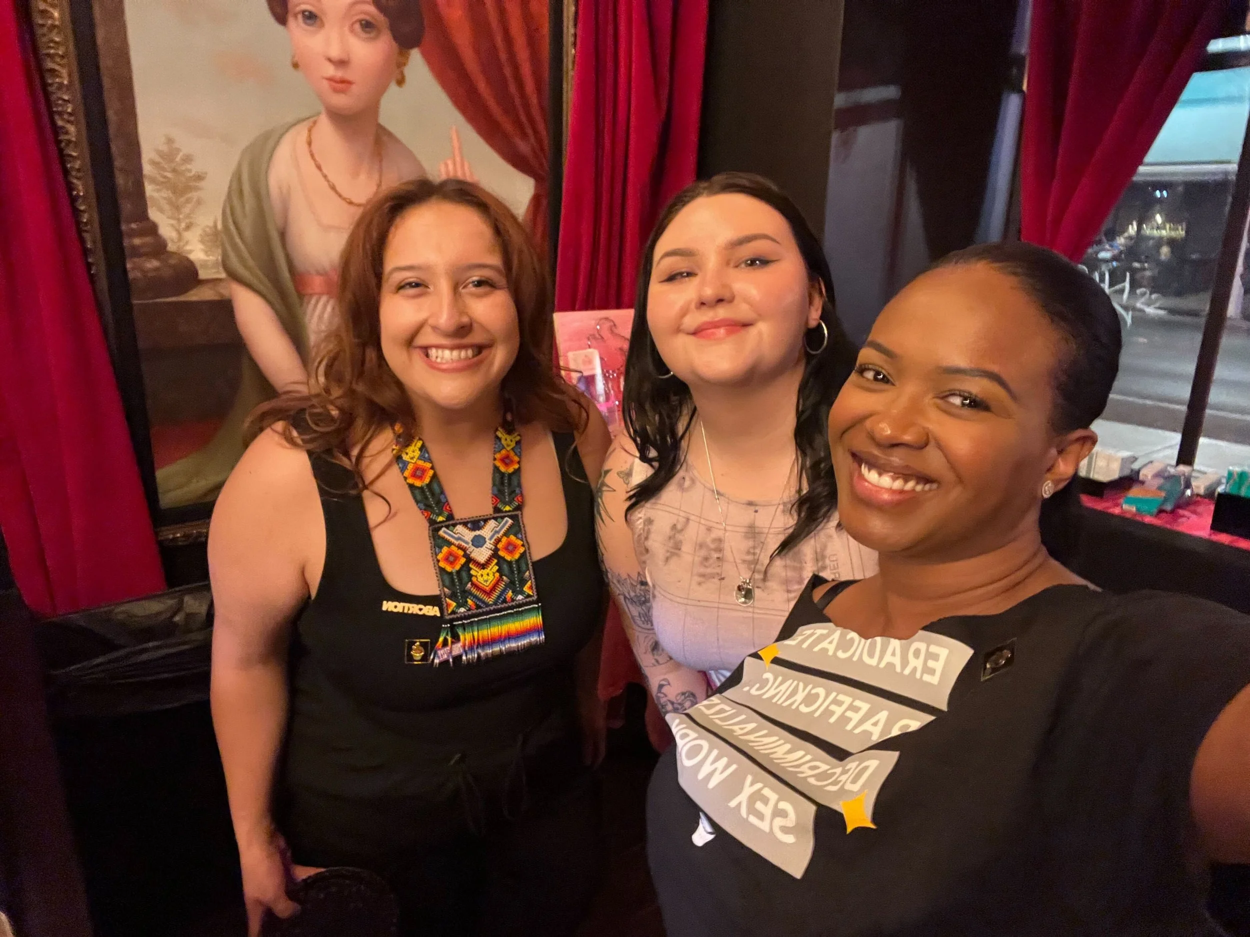 Three femme presenting individuals at a celebration event with The Cupcake Girls. Red curtains and neon signs in the background.