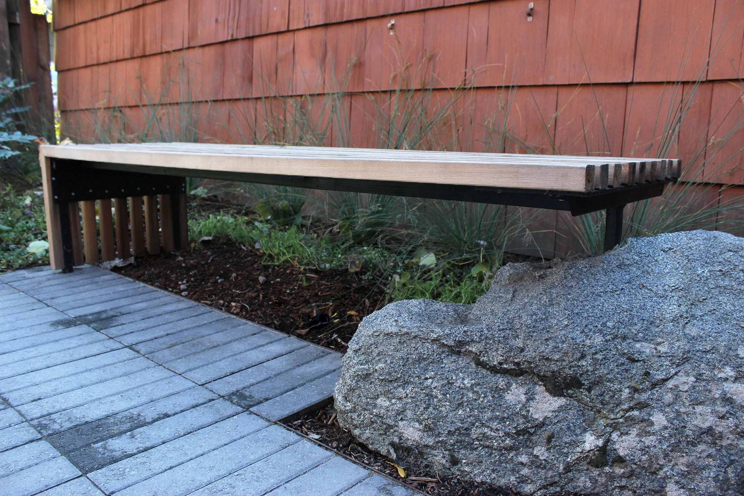 a wood bench integrated into a boulder for seating, with grasses and pavers nearby