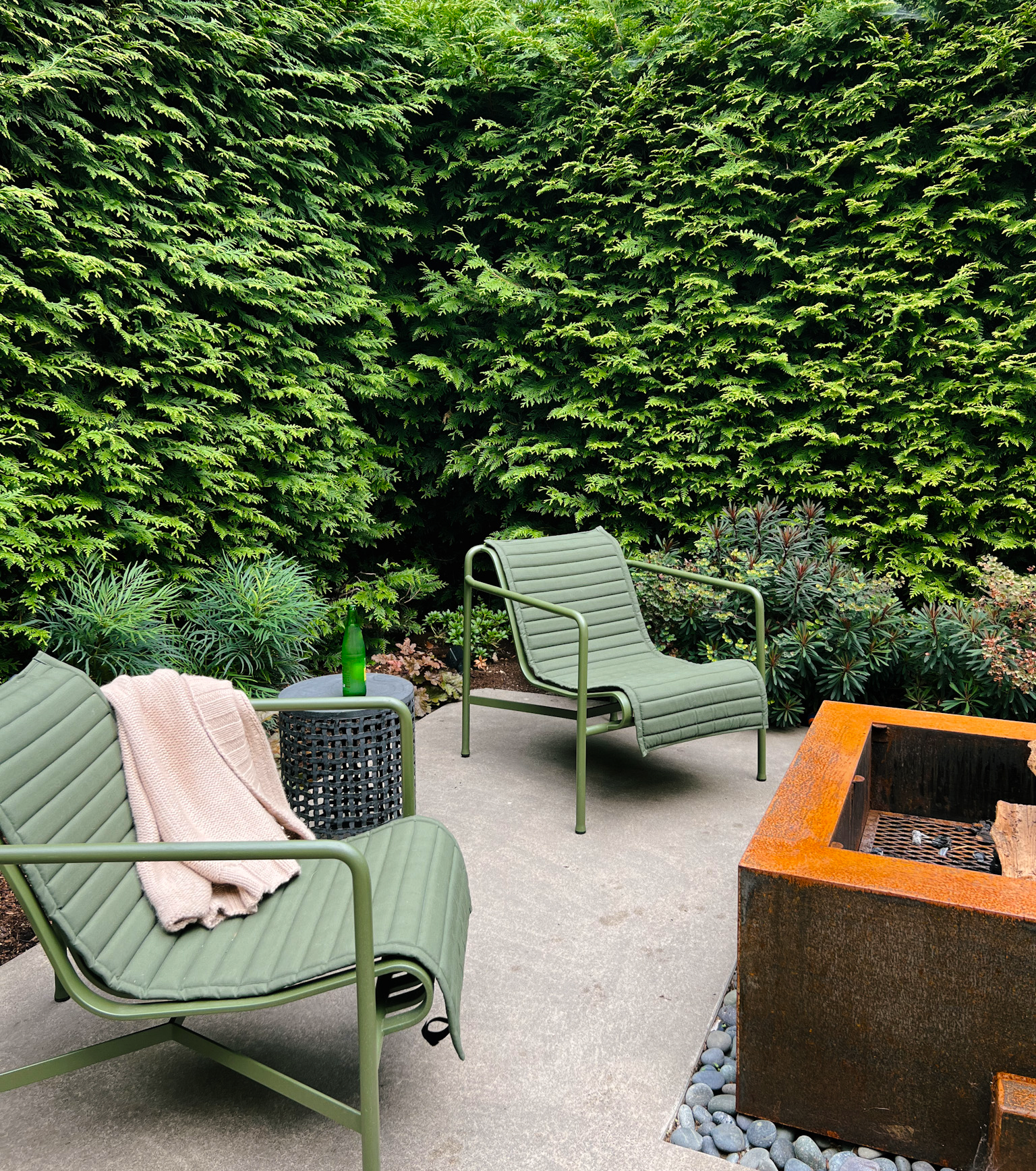 Two green chairs in front of a corten steel firepit surrounded by tall privacy hedges