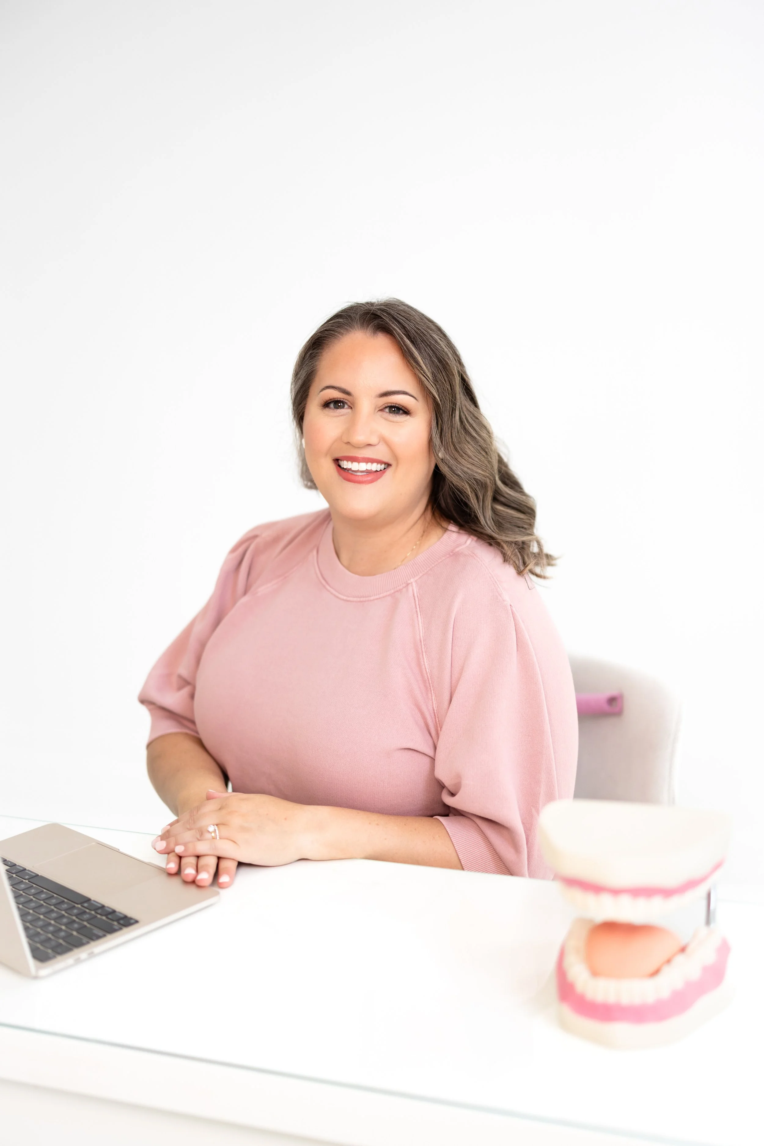 A smiling woman with wavy hair sitting at a white desk with a dental model in front of her and a laptop to her side.