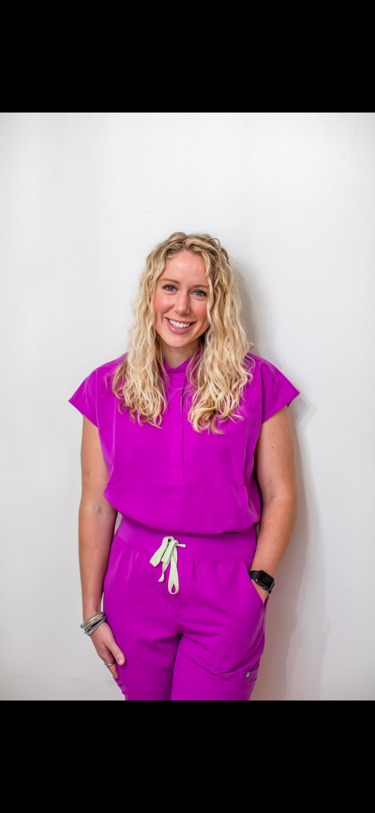 A smiling woman with blonde curly hair wearing a bright purple medical scrubs outfit and a smartwatch, standing against a white wall.